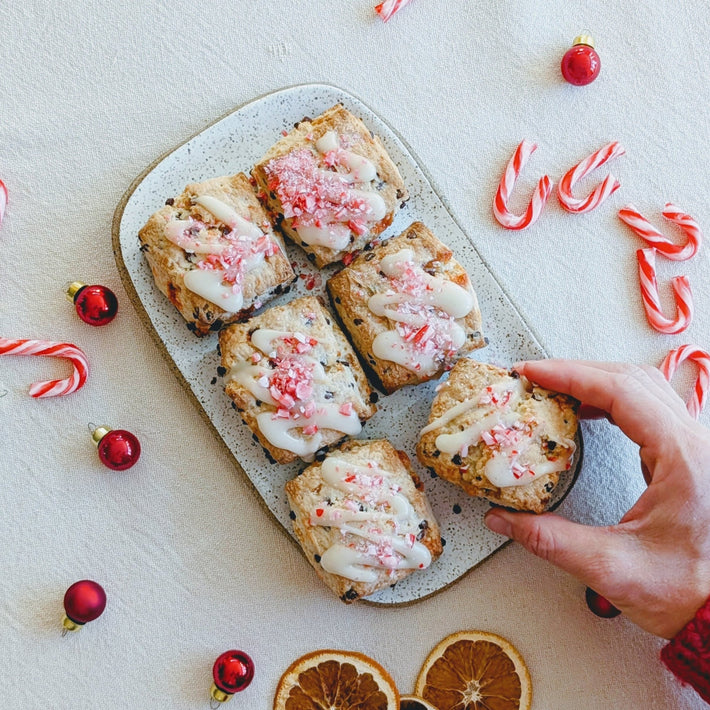 A hand reaching for a plate of iced baked goods with crushed candy canes, surrounded by holiday decorations.