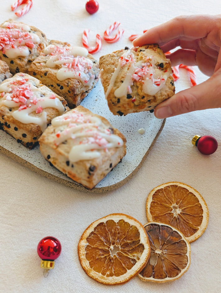 A hand reaching for a scone topped with icing and crushed candy canes, with holiday decorations and dried orange slices.
