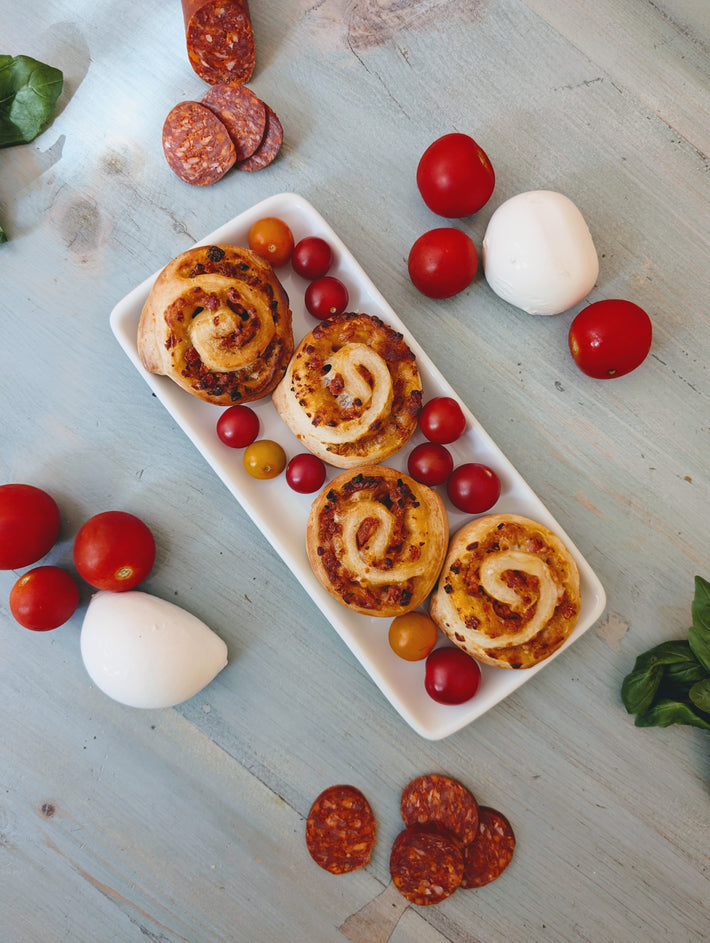 Four pinwheel pastries on a white plate with cherry tomatoes, mozzarella and sliced salami on a pale wood table.