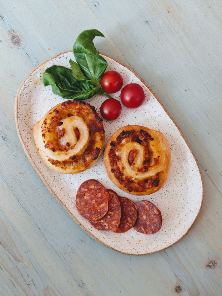 Two savory pinwheel pastries with sliced chorizo, cherry tomatoes, and basil on a speckled oval plate on pale blue wood