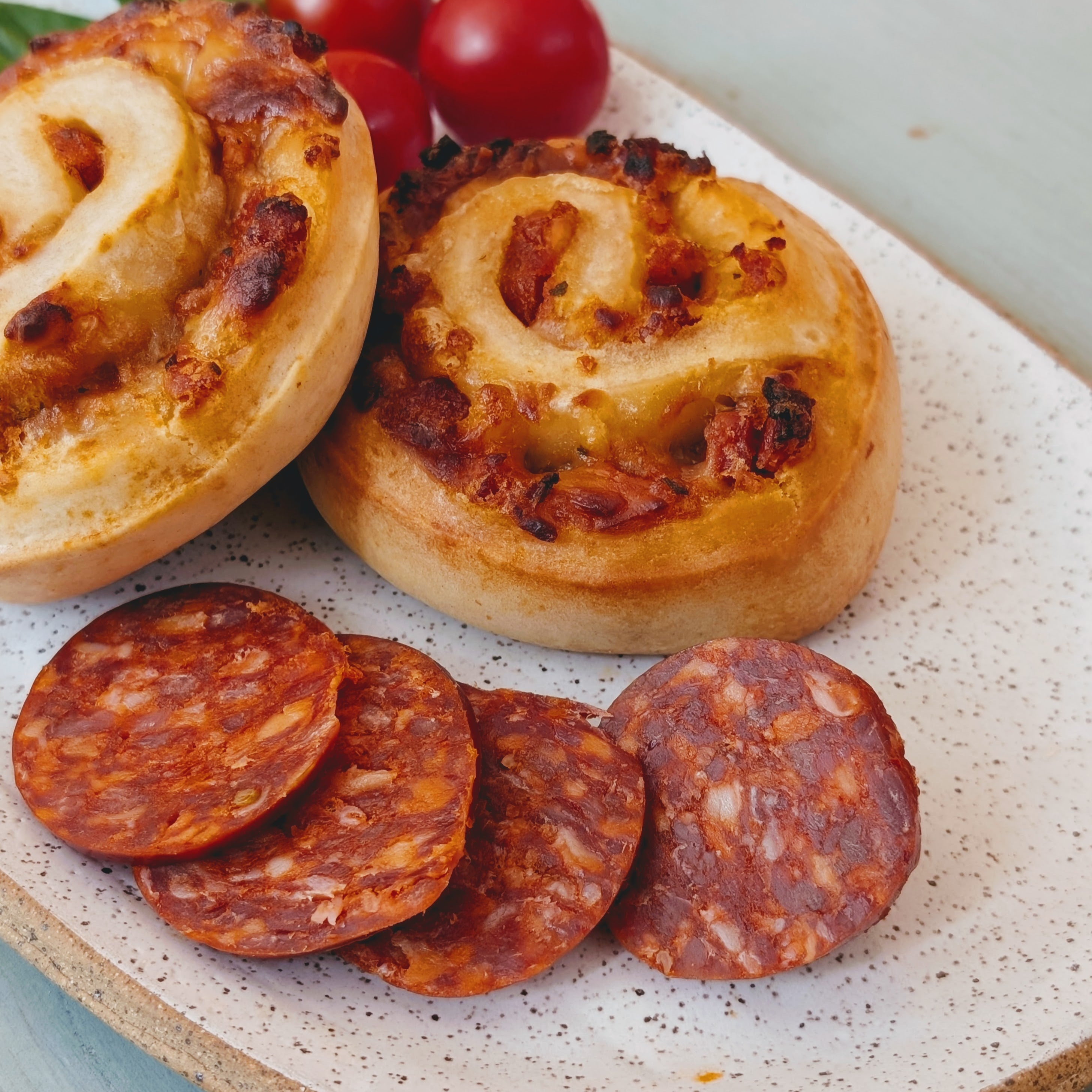 Two savory cheese-and-bacon pinwheel pastries on a speckled plate with sliced chorizo rounds and cherry tomatoes.
