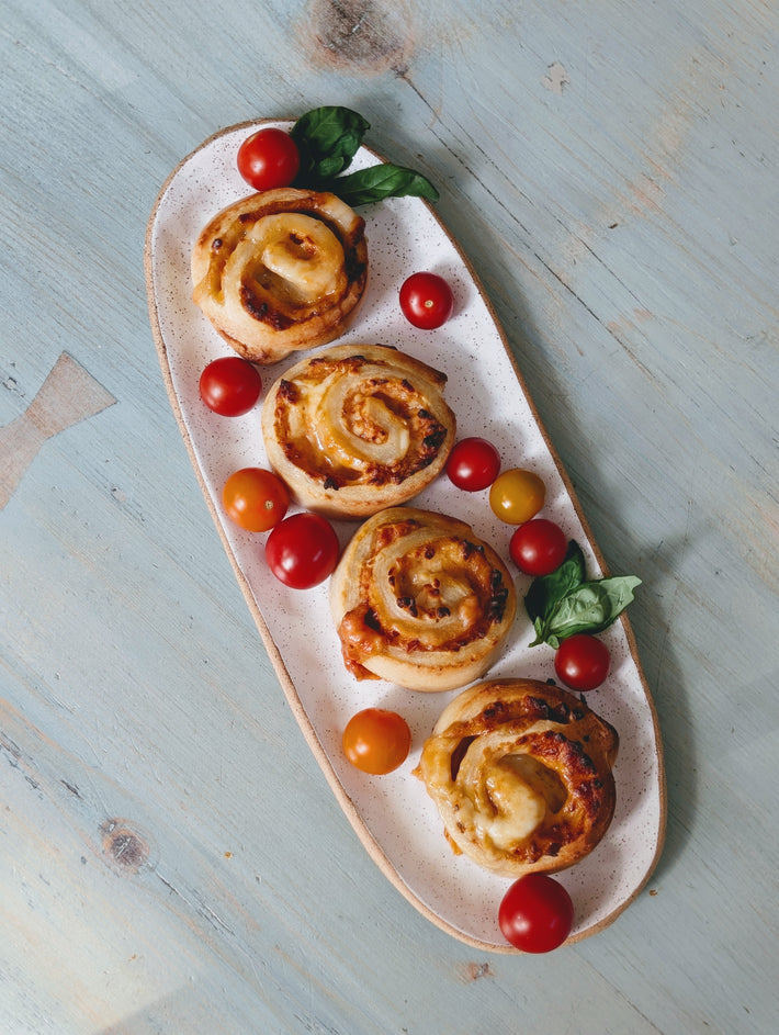 Four savory cheese-tomato pinwheel pastries with basil and cherry tomatoes on an oval speckled plate on a blue wooden table.