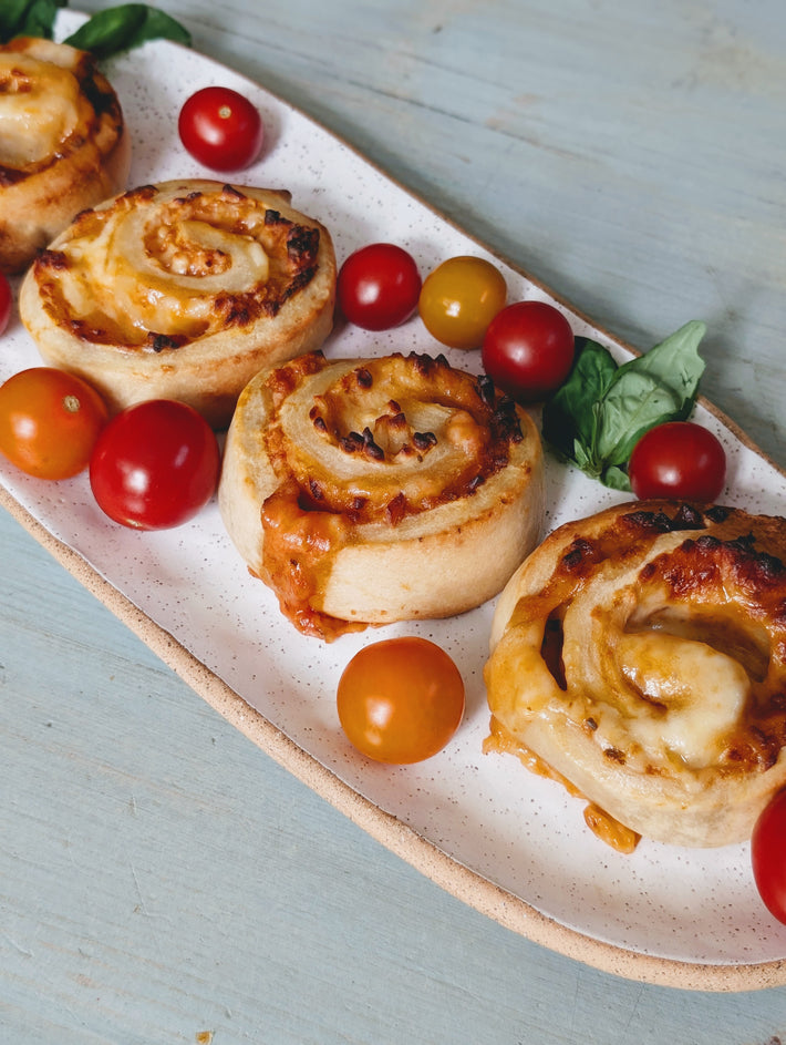 Baked cheesy pizza pinwheels on a speckled white platter with red and yellow cherry tomatoes and basil on a pale blue table.