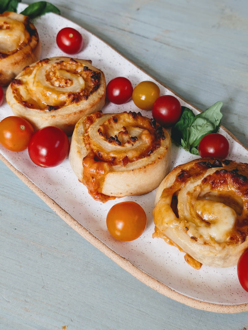 Baked cheesy pizza pinwheels on a speckled white platter with red and yellow cherry tomatoes and basil on a pale blue table.