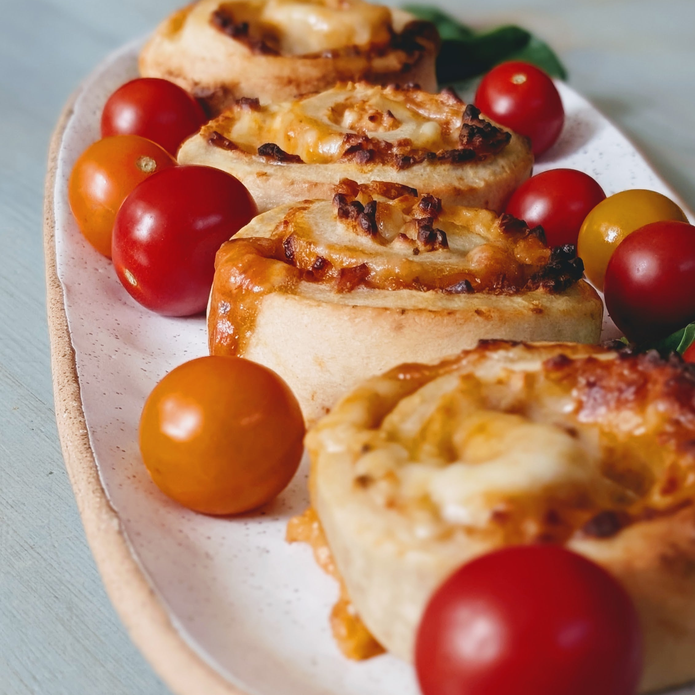 Cheesy pizza pinwheel rolls with browned edges arranged on an oval speckled platter with red and yellow cherry tomatoes