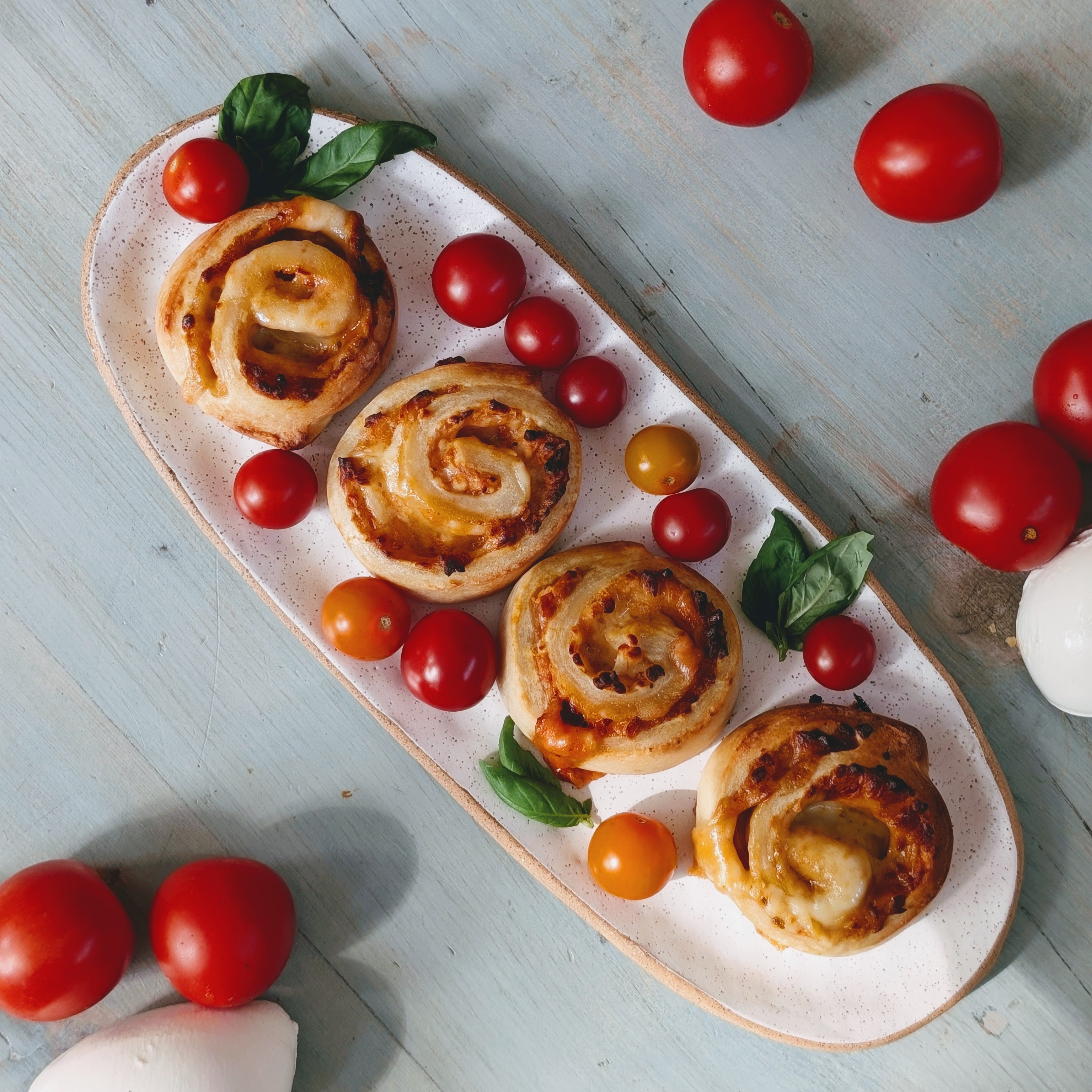 Four cheese pinwheel pastries with cherry tomatoes and basil on a speckled oval platter on a pale blue wooden table.