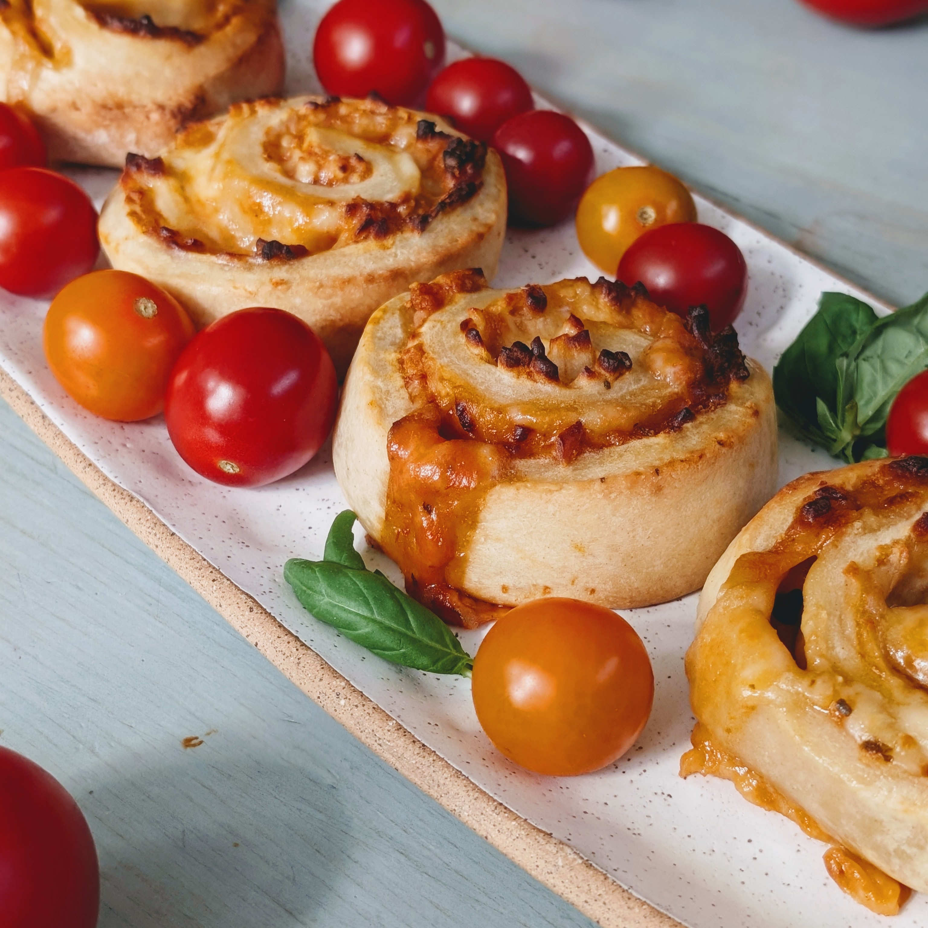 Baked cheesy pinwheel pastries on a rectangular platter, garnished with red and orange cherry tomatoes and basil leaves.