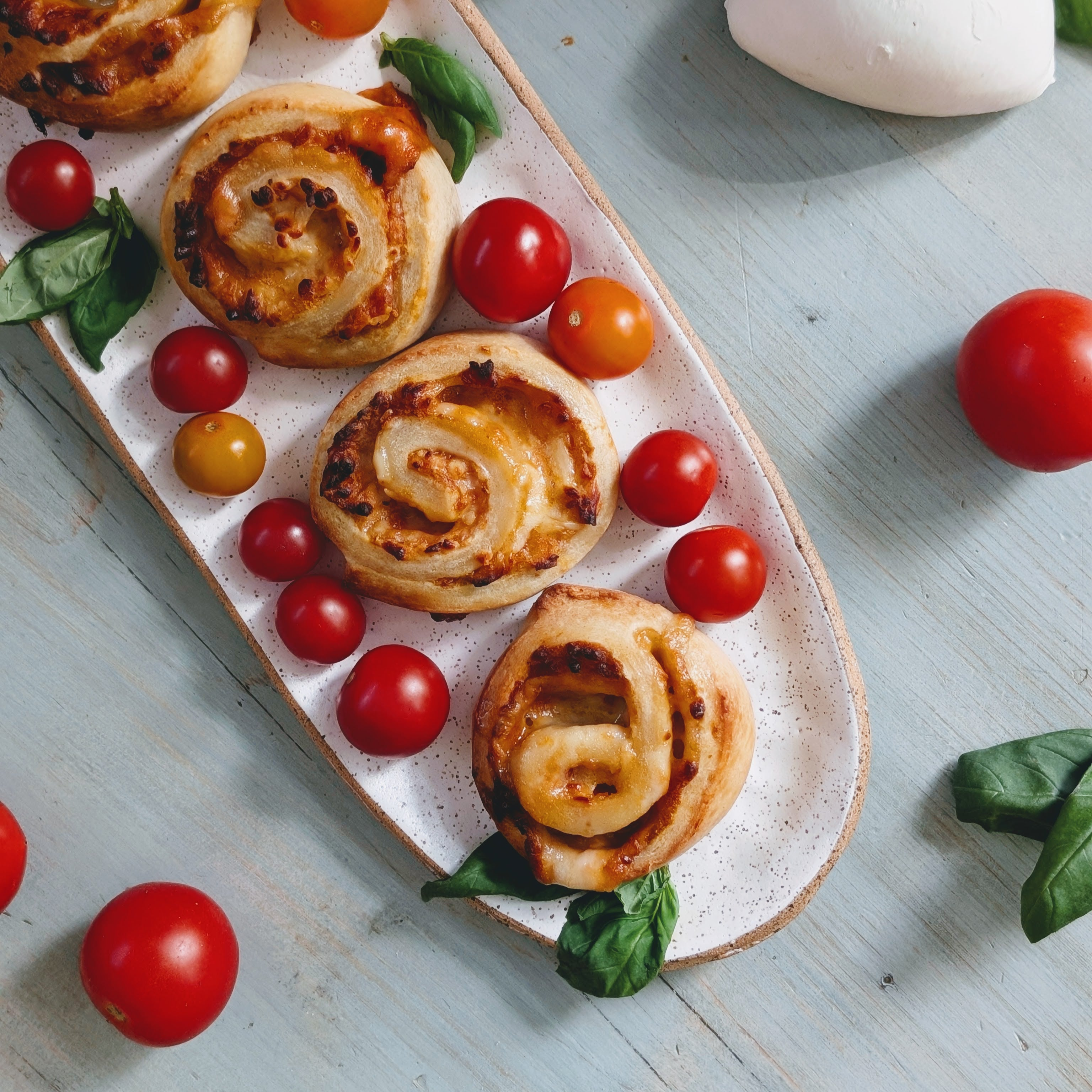 Three savory cheese-and-tomato pinwheel rolls on a speckled oval platter with cherry tomatoes and basil leaves