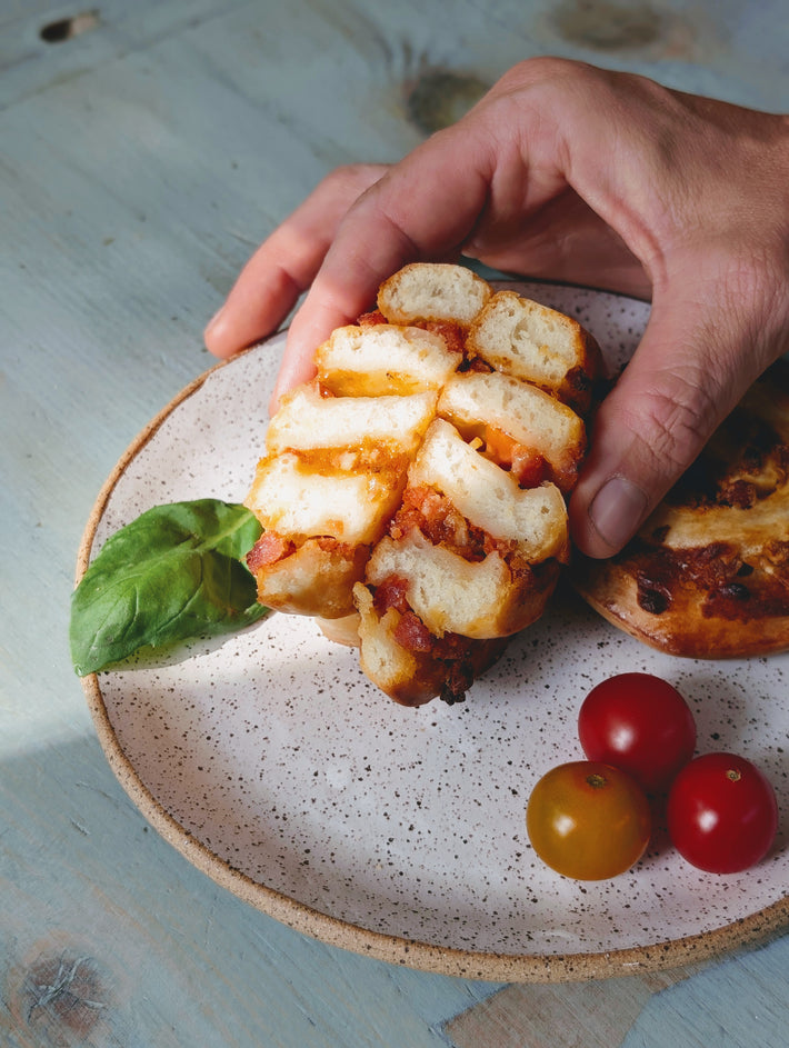Hand holding a pulled-apart cheesy bread piece on a speckled plate with a basil leaf and three cherry tomatoes.