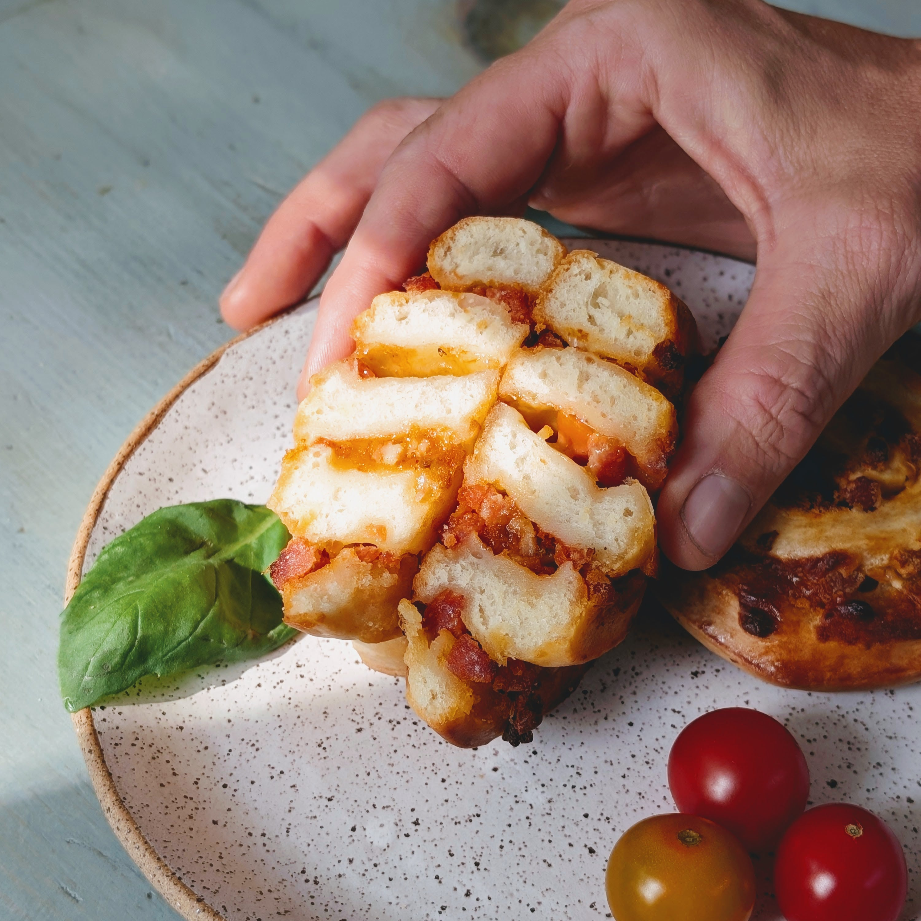 Hand holding sliced toasted bread with melted cheese and tomato, on a speckled plate with basil and cherry tomatoes.