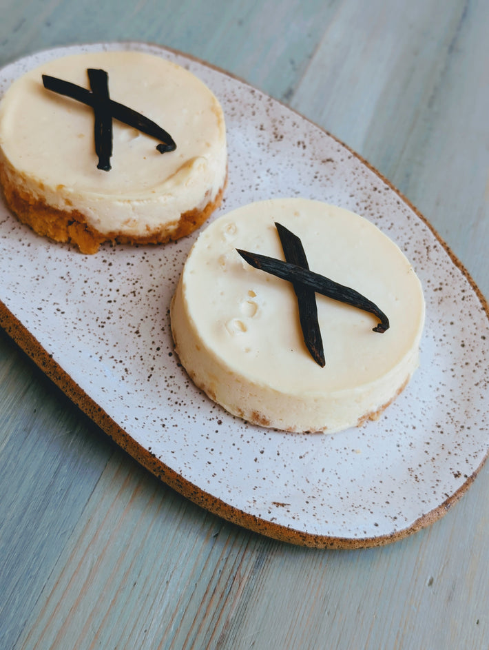 Two small plain cheesecakes on a speckled platter, each topped with crossed vanilla bean pieces