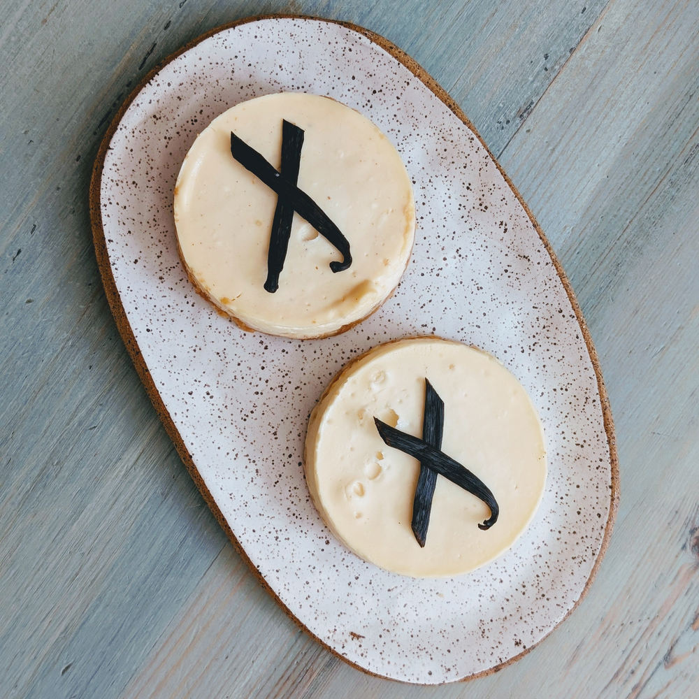 Two small round cheesecakes with cream topping and black chocolate X decorations on a speckled oval plate.