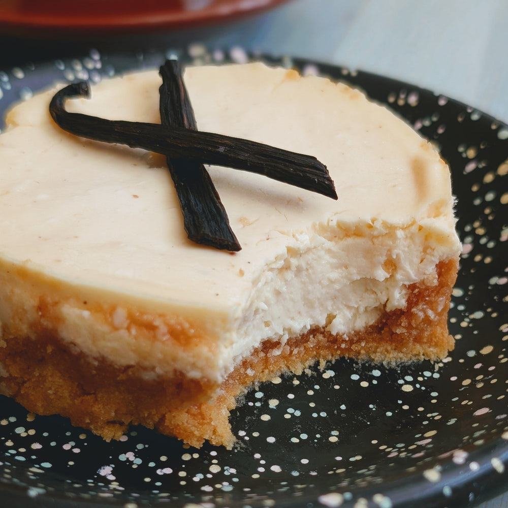 Cheesecake slice with graham crust and two vanilla bean pods on top, a bite taken, on a speckled black plate.