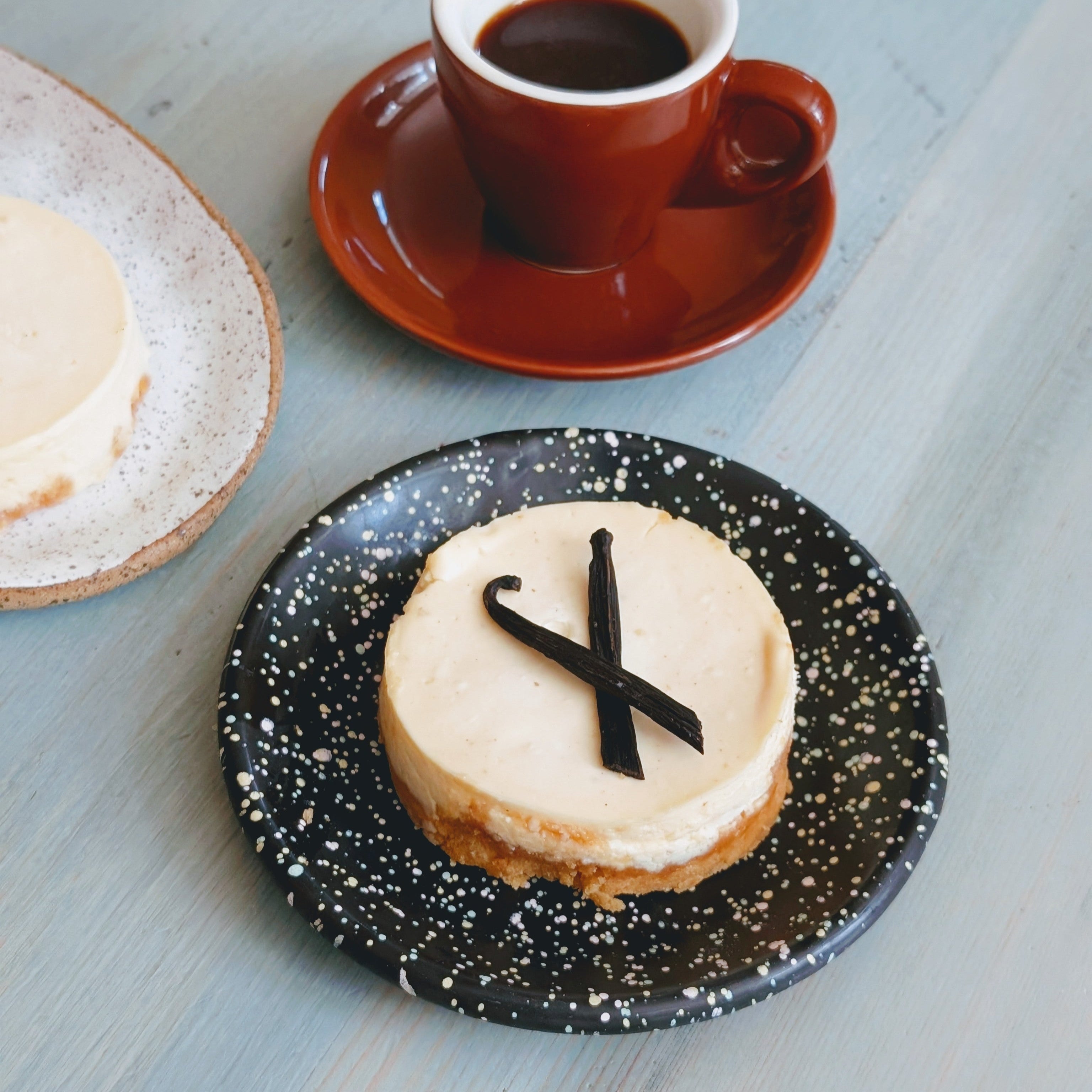Mini vanilla cheesecake topped with two crossed dark chocolate sticks on a speckled black plate beside an espresso cup.