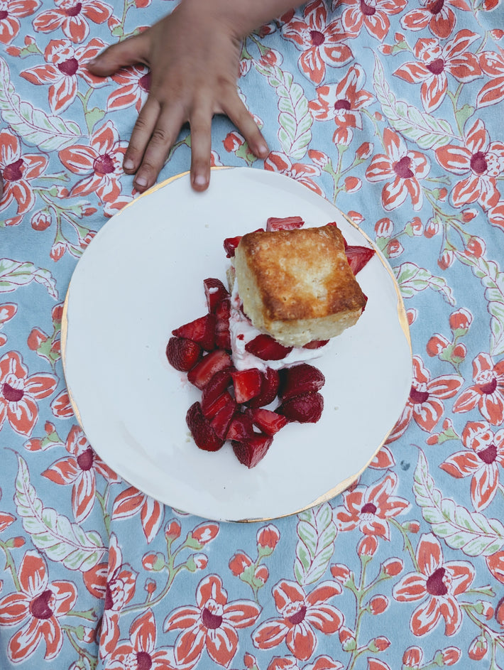 White plate with strawberry shortcake and chopped strawberries on a floral tablecloth, a child's hand reaching from top.