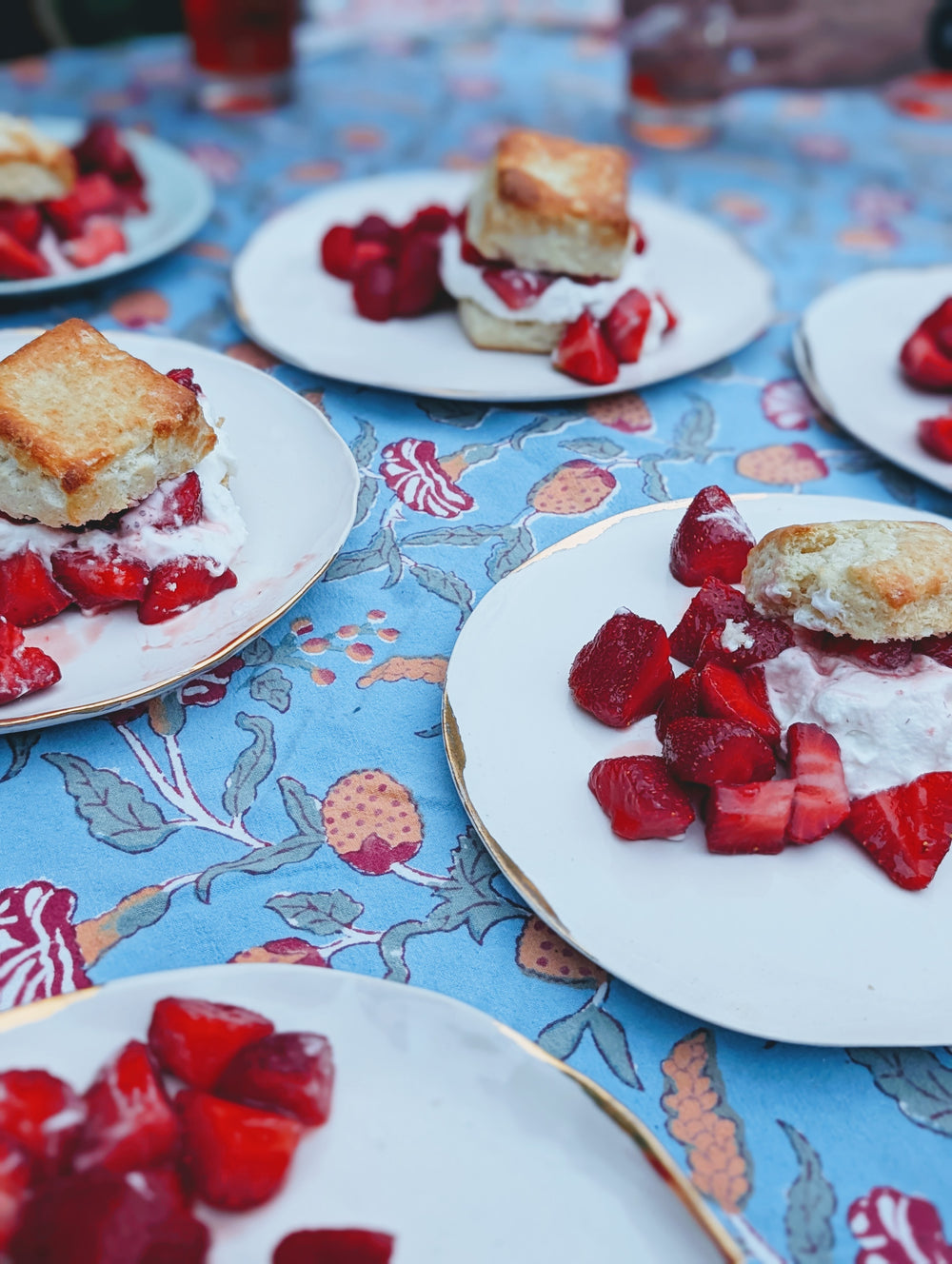 Plates of strawberry shortcakes with buttery biscuits, whipped cream and sliced strawberries on a blue floral tablecloth