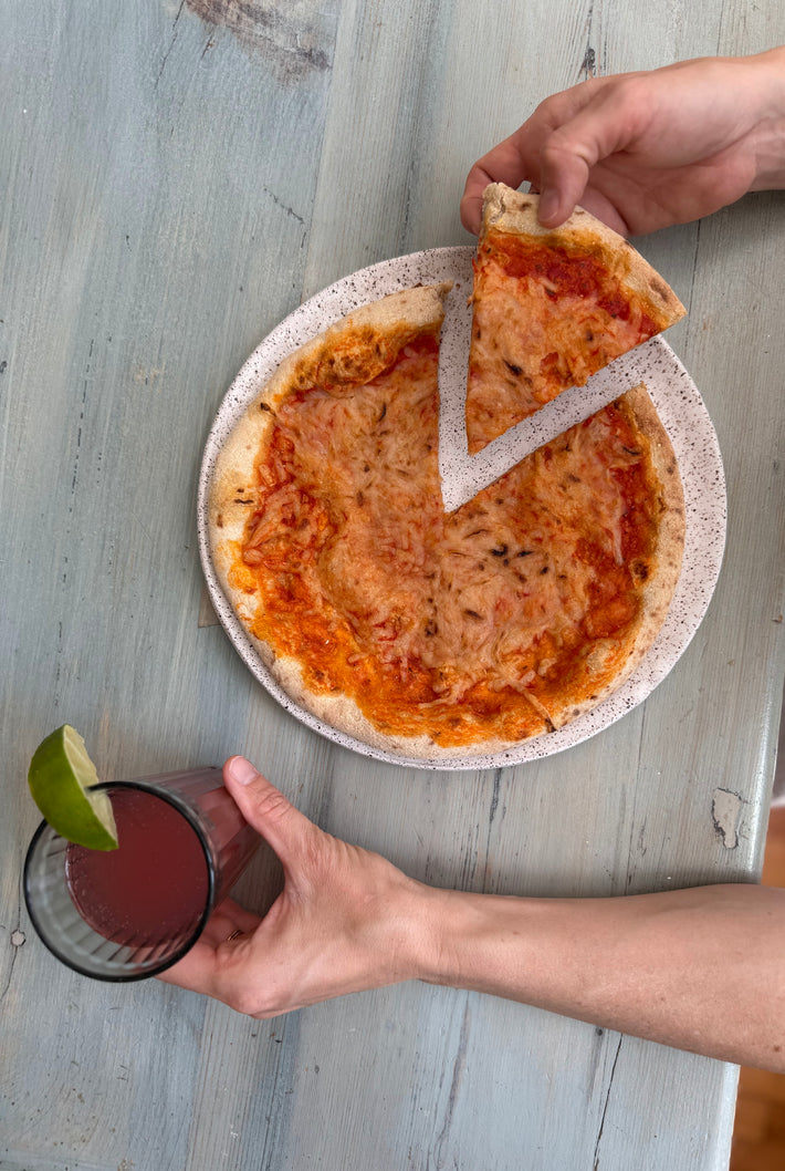 A hand holds a slice of Wildgrain Plant-Based Cheese Pizza from a round plate, while another hand holds a glass with a drink and a lime wedge.