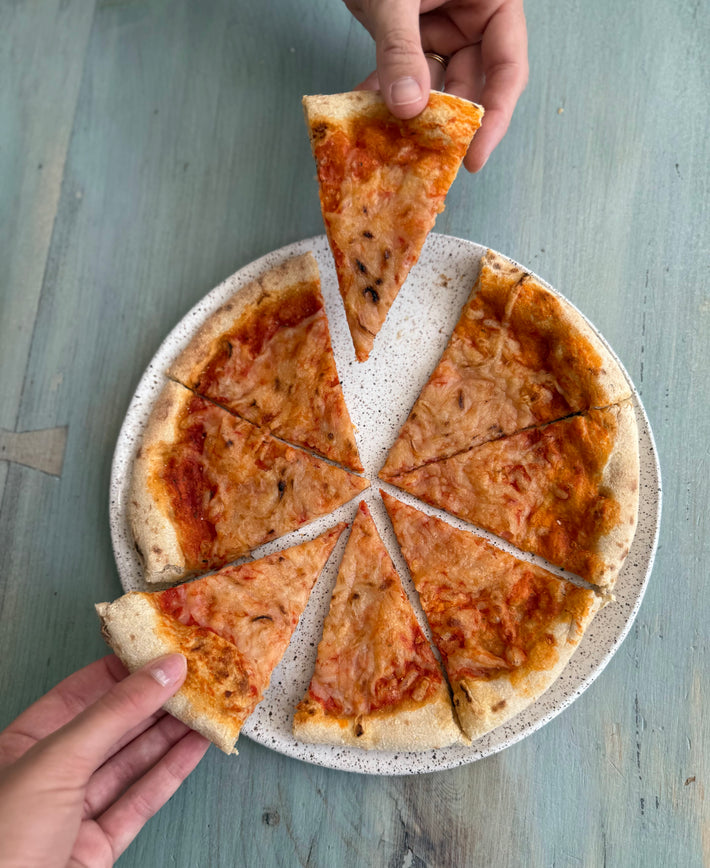 Wildgrain Plant-Based Cheese Pizza on a speckled plate with several slices cut, one slice being lifted by a hand.