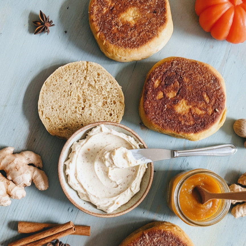 Toasted English muffins with spiced whipped butter and pumpkin spread, cinnamon sticks and fresh ginger on a pale blue board.