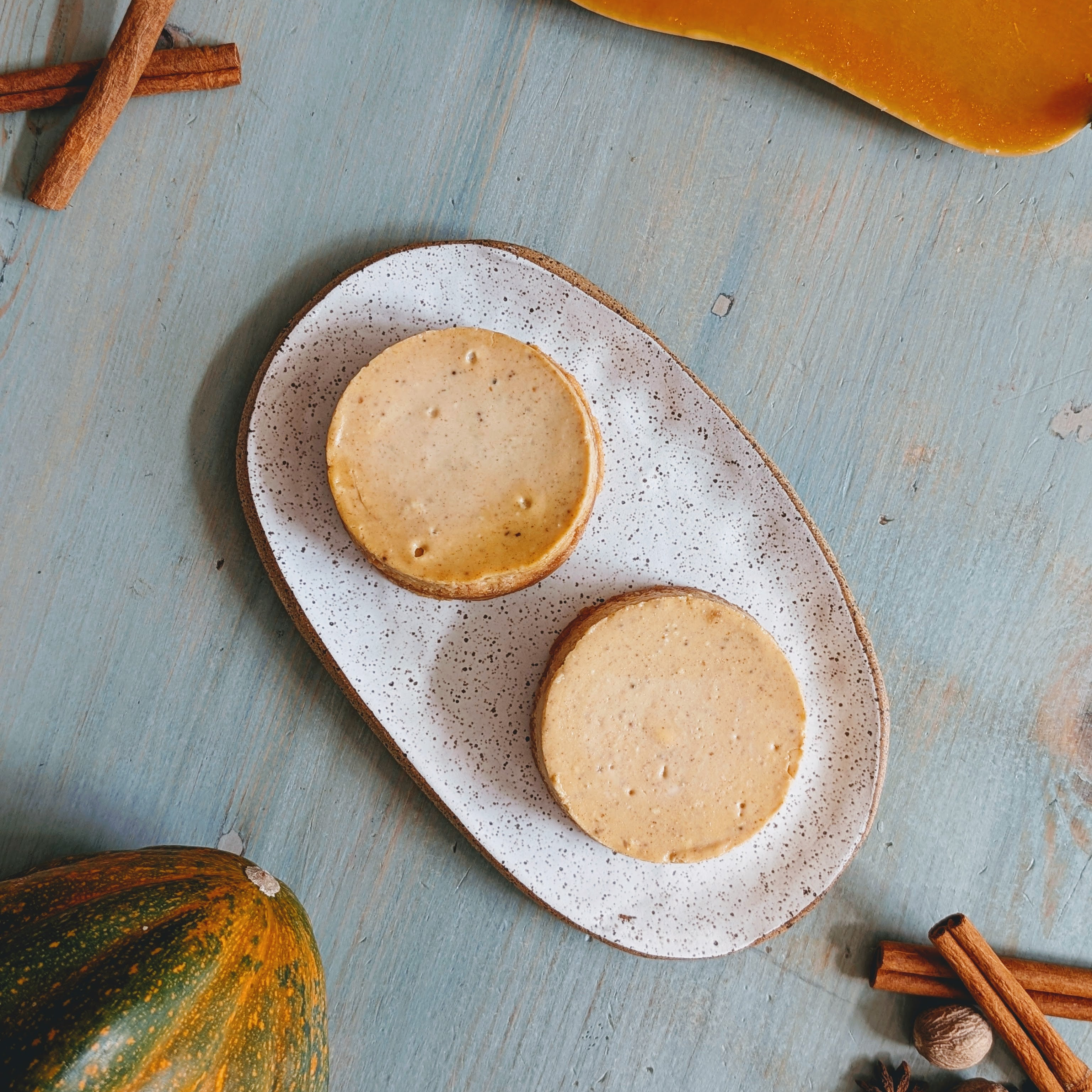 Two round beige tarts on a speckled oval plate, cinnamon sticks and a small pumpkin on a blue wooden table.