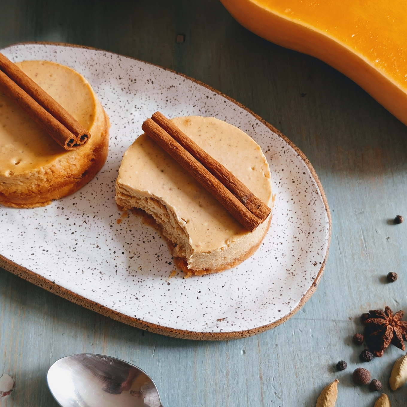 Two mini pumpkin cheesecakes on speckled plate, one with a bite and cinnamon sticks on top; spoon and spices nearby.