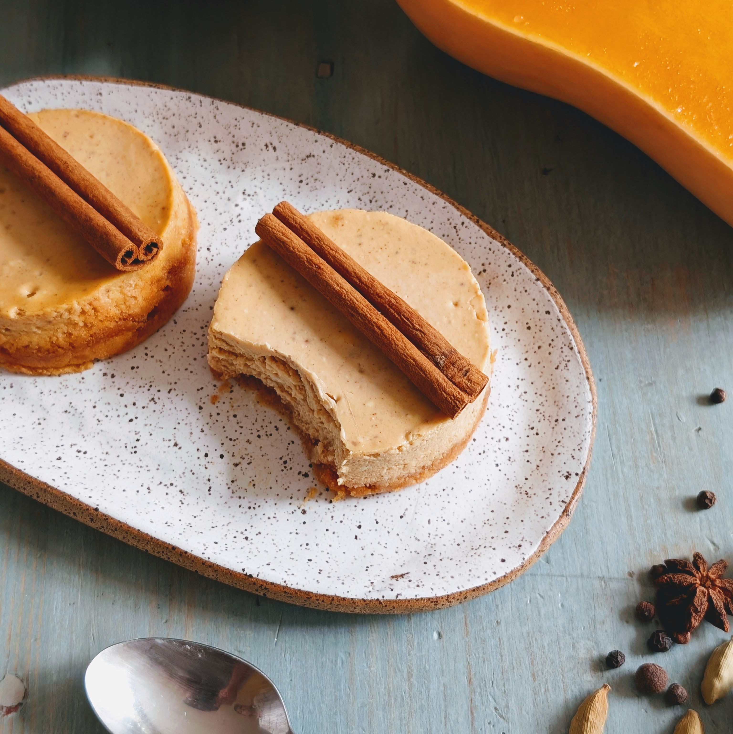 Two mini pumpkin cheesecakes on speckled plate, one with a bite and cinnamon sticks on top; spoon and spices nearby.
