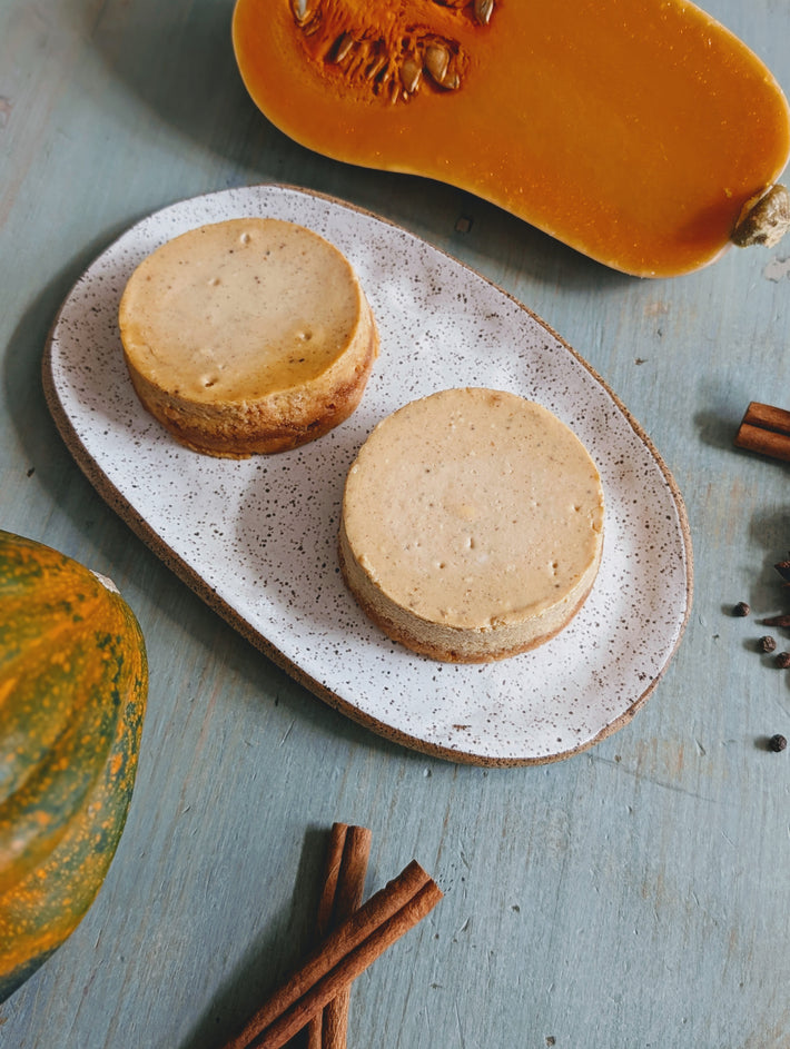 Two mini pumpkin cheesecakes on a speckled oval plate with cinnamon sticks and halved squash on blue wood