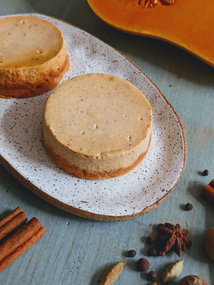 Two mini pumpkin cheesecakes on a speckled oval plate with cinnamon sticks, star anise and cardamom on a blue wooden surface.