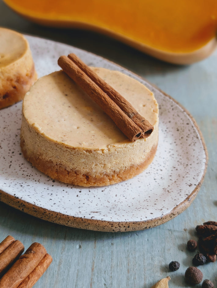 Spiced mini cheesecake topped with a cinnamon stick on a speckled plate, whole spices scattered nearby