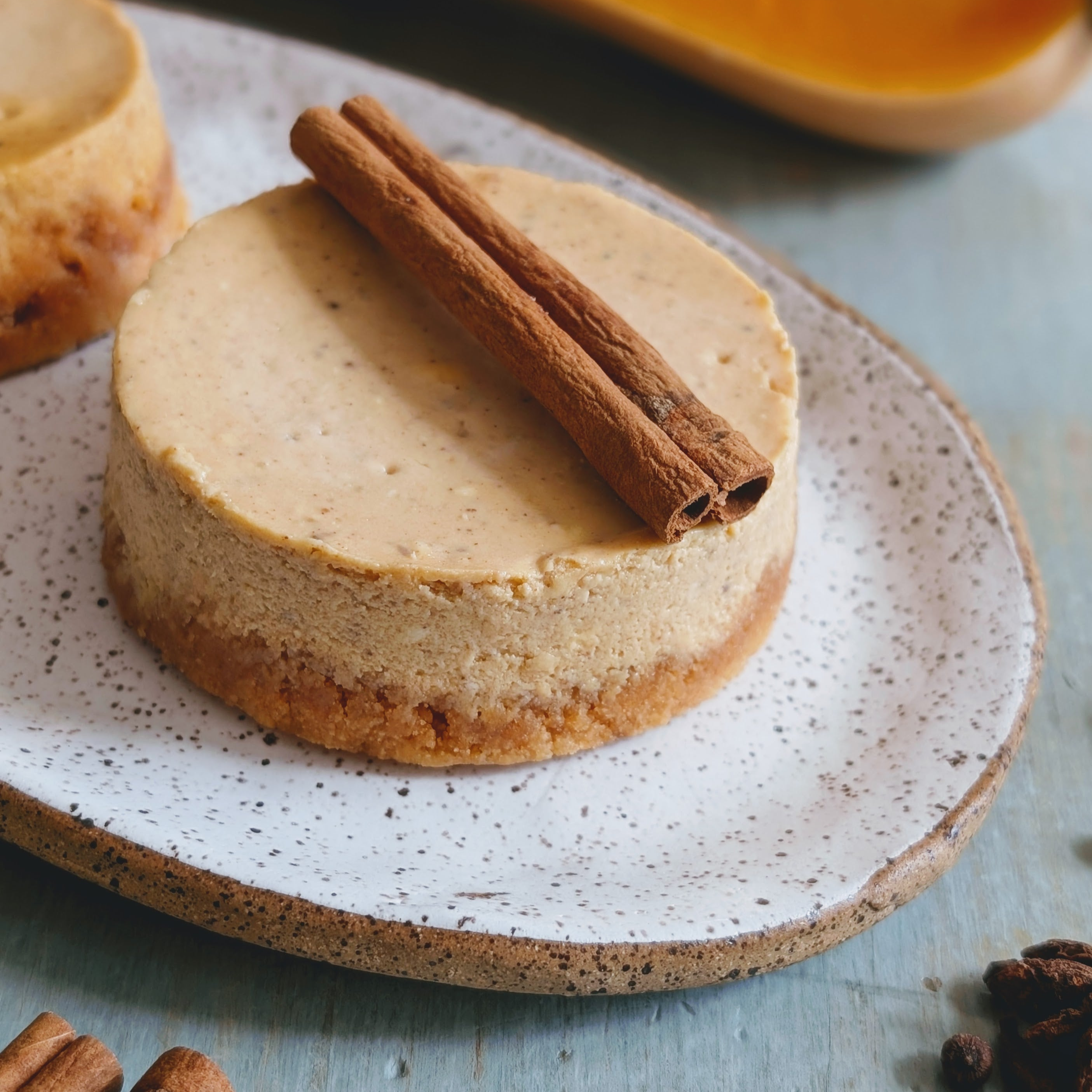 Mini spiced cheesecake with a cinnamon stick garnish on a speckled ceramic plate.