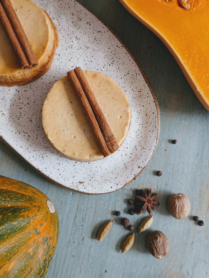 Two mini pumpkin cheesecakes on a speckled plate with cinnamon sticks, plus pumpkin, star anise, cardamom, and nutmeg.