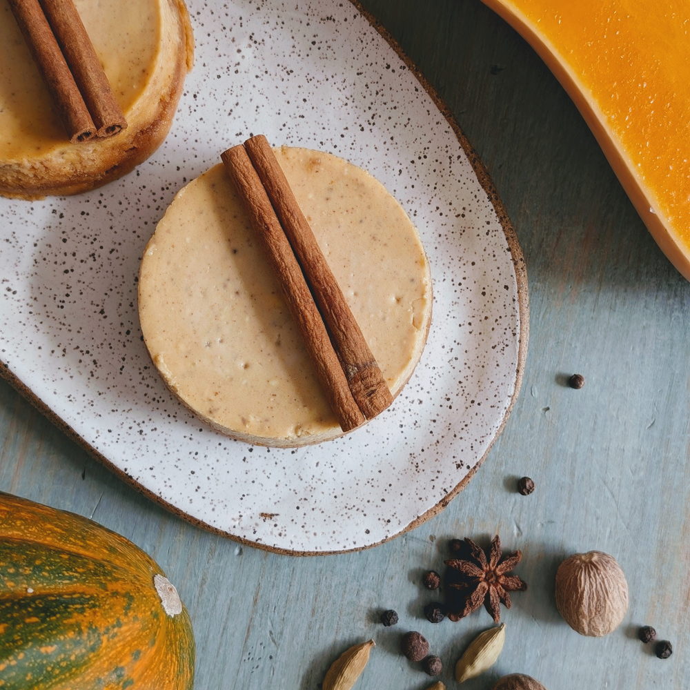 Two mini pumpkin-spice cheesecakes on a speckled plate garnished with cinnamon sticks, with pumpkin and whole spices nearby.