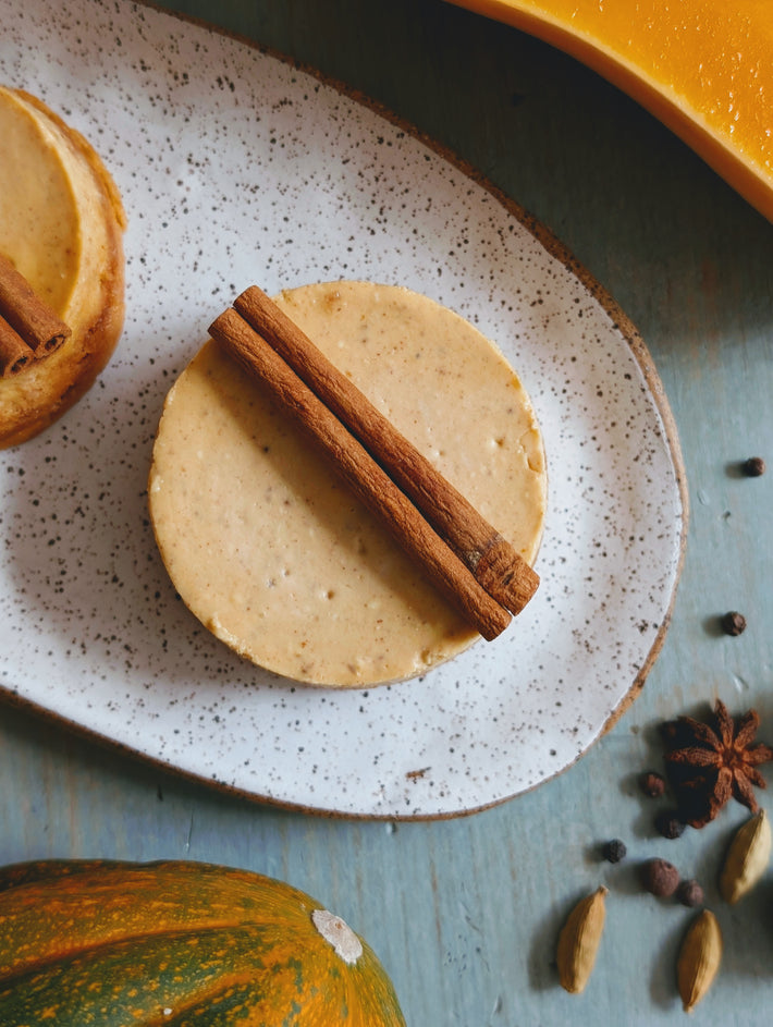 Round spiced pumpkin cheesecake topped with two cinnamon sticks on speckled white plate, with star anise and cardamom nearby.