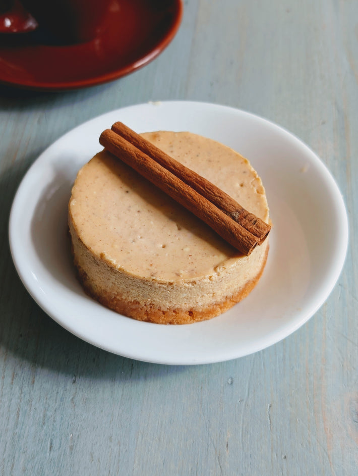 Mini cinnamon cheesecake on a white saucer topped with two cinnamon sticks, on a light blue wooden table.