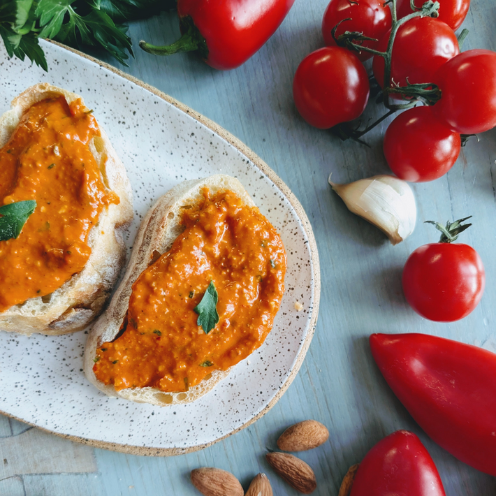 Two slices of crusty bread with red pepper spread on a speckled plate, surrounded by cherry tomatoes, garlic and almonds.