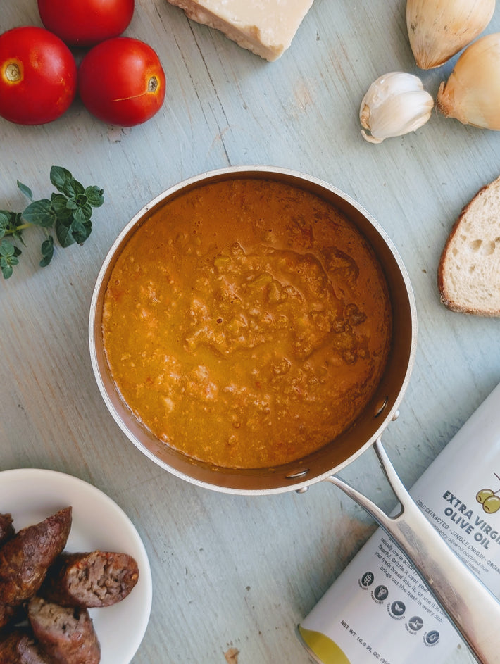 Saucepan with red sauce on a wooden surface with tomatoes, bread, and herbs.
