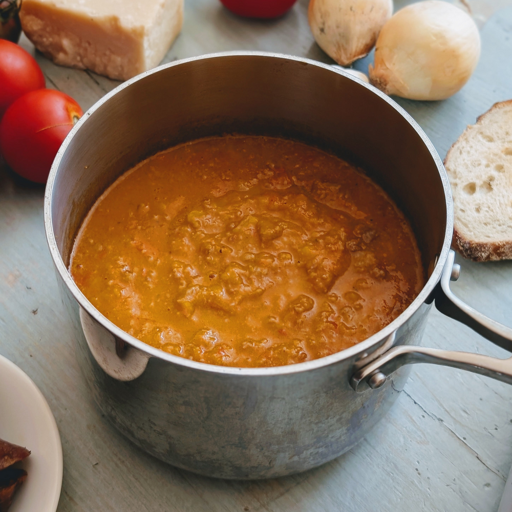Pot of tomato soup in a saucepan on a table with tomatoes, onions, a slice of bread, and a wedge of cheese.