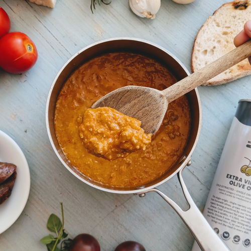 Top-down view of a saucepan of thick tomato sauce being stirred with a wooden spoon; bottle labeled EXTRA VIRGIN OLIVE OIL visible.