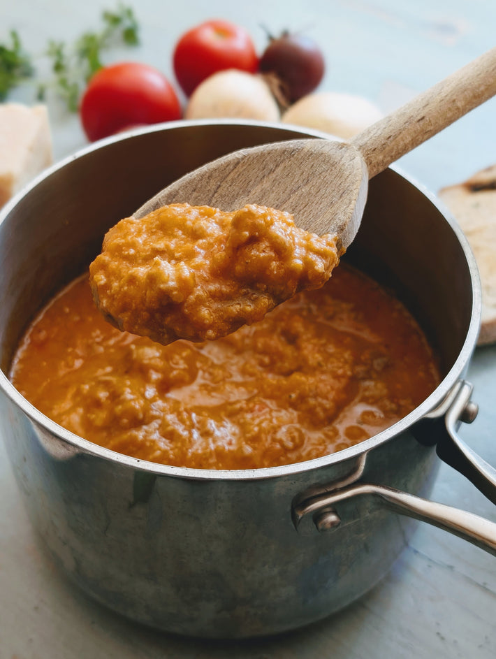 Serving of tomato-based Sausage Ragu Sauce with a wooden spoon in a pot, surrounded by tomatoes and onions.