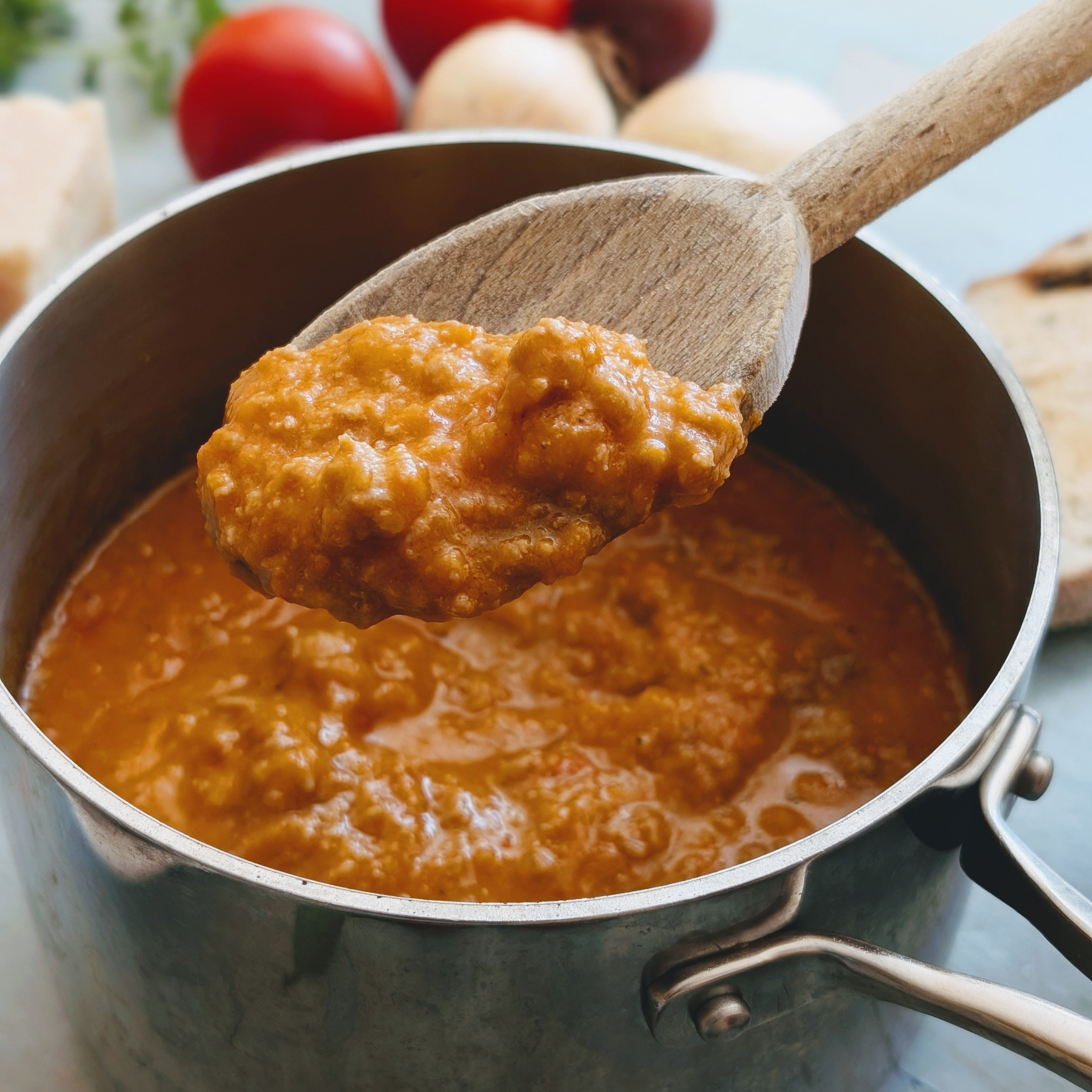 Wooden spoon lifting chunky tomato sauce from a saucepan with tomatoes and garlic blurred in the background.