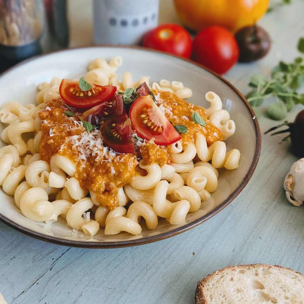 Bowl of curly pasta topped with sausage ragu, cherry tomato halves and grated cheese on a light blue table with a slice of bread and fresh herbs
