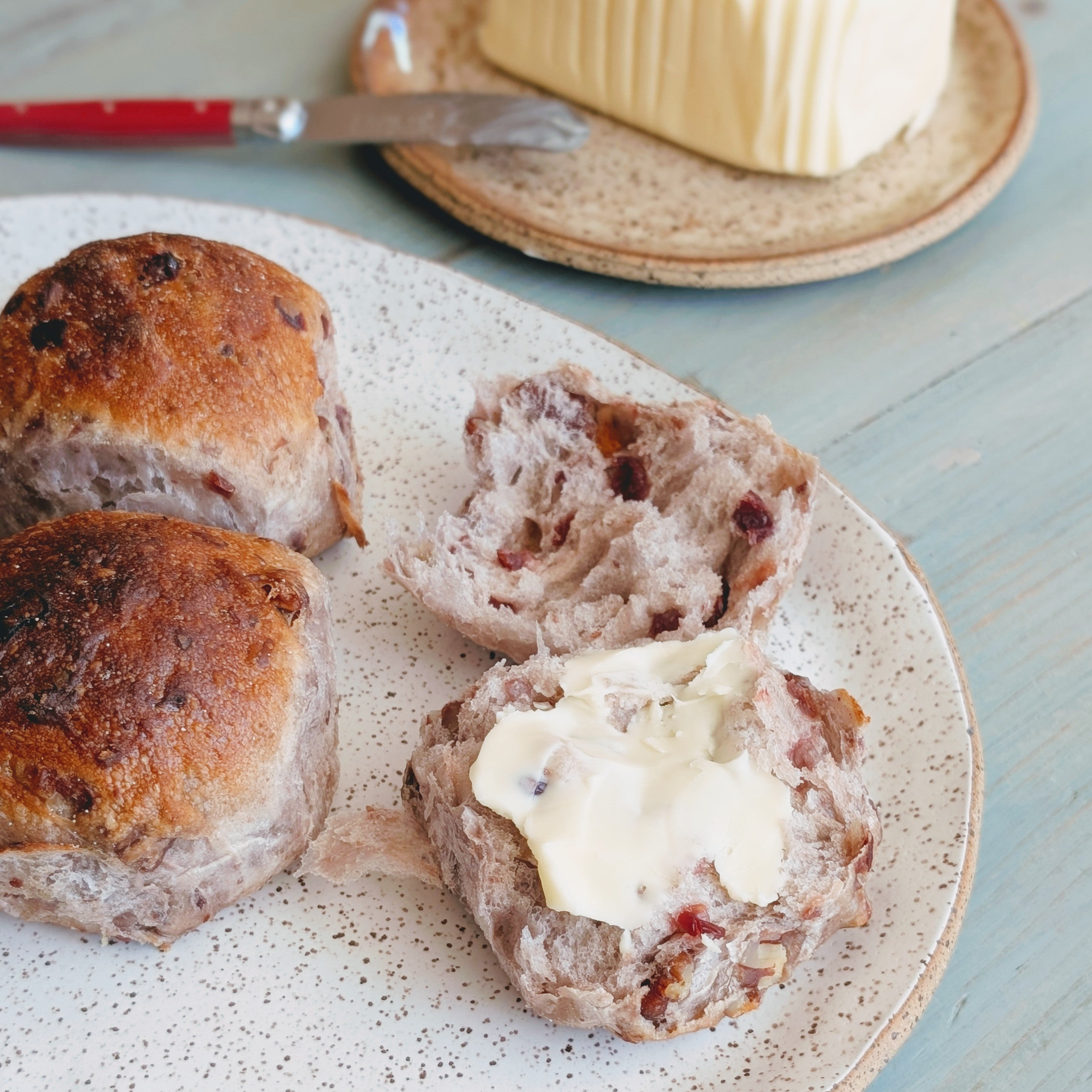 Split cranberry and orange tea buns on speckled plate, one with softened butter; butter dish and knife in background.