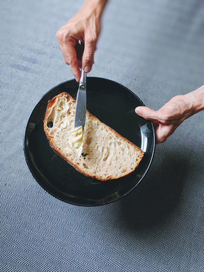 Hands holding a black plate with a slice of crusty bread being buttered with a knife.
