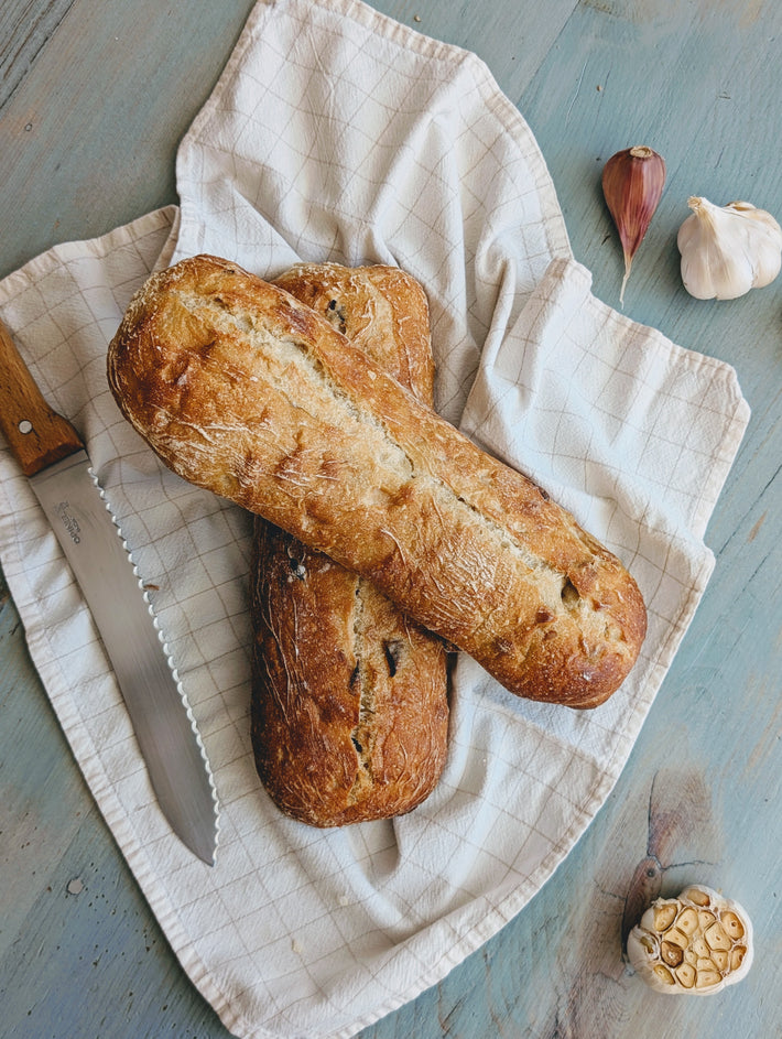 Two loaves of Slow-Fermented Garlic Demi Ciabattas on a white cloth with garlic and shallots nearby.