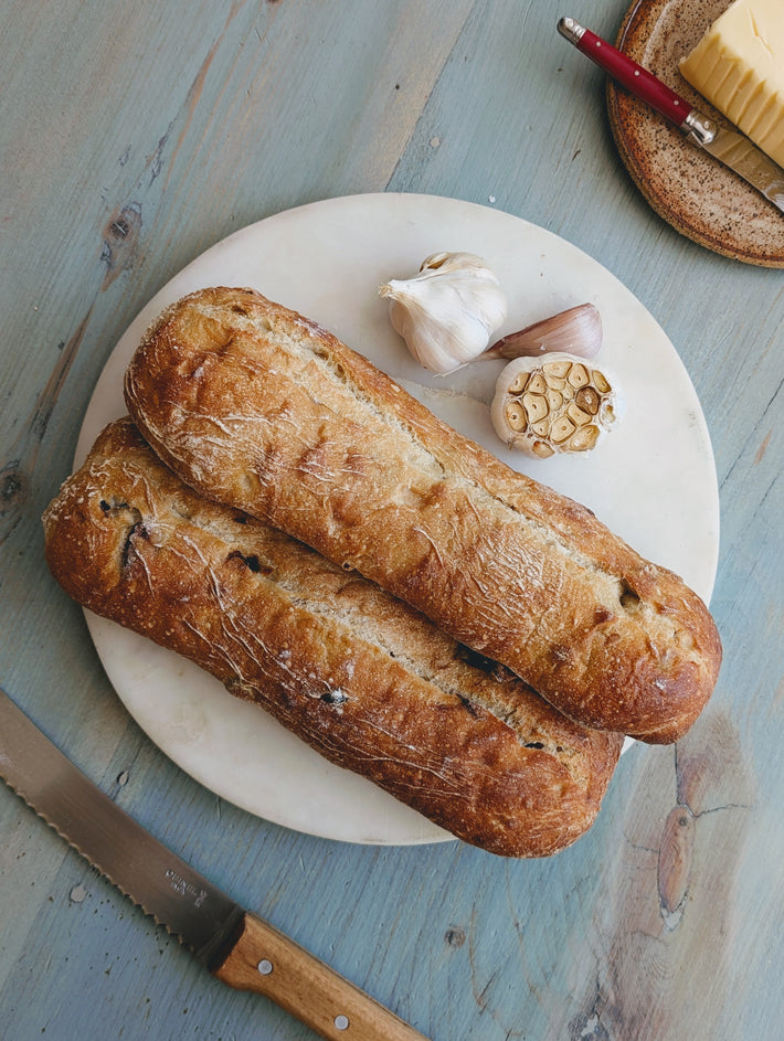 Two loaves of Slow-Fermented Garlic Demi Ciabattas on a marble board with garlic cloves and a knife.