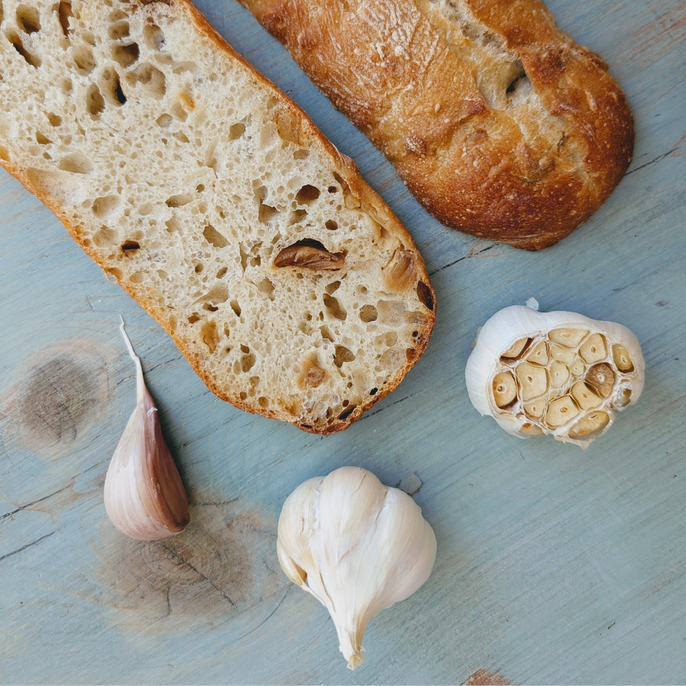 Two slices of crusty bread with a halved garlic head, a whole bulb and a clove on a blue wooden board