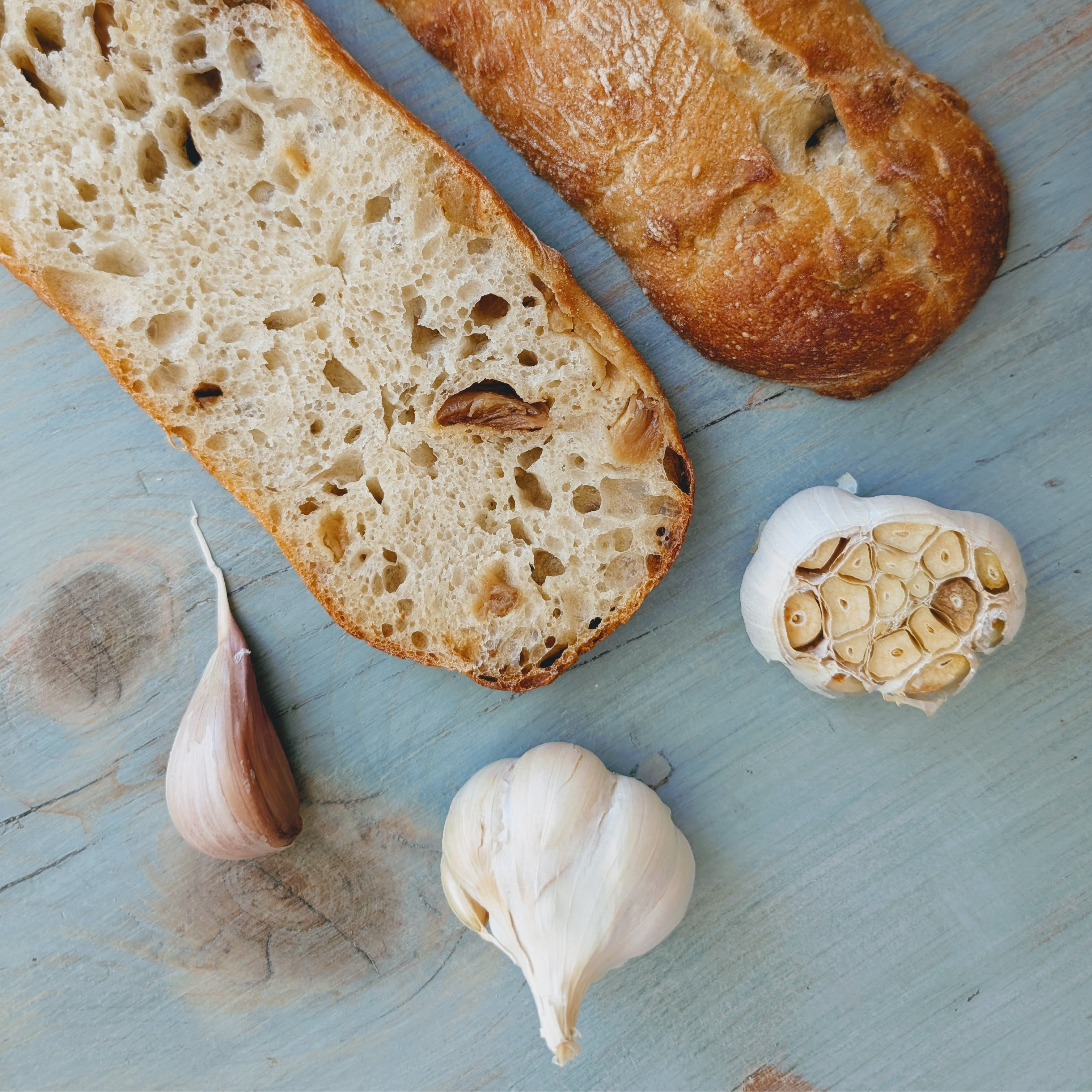 Two slices of crusty bread with a halved garlic head, a whole bulb and a clove on a blue wooden board