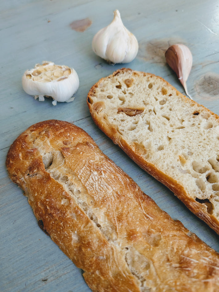 Sliced Slow-Fermented Garlic Demi Ciabattas on a blue surface with garlic cloves.
