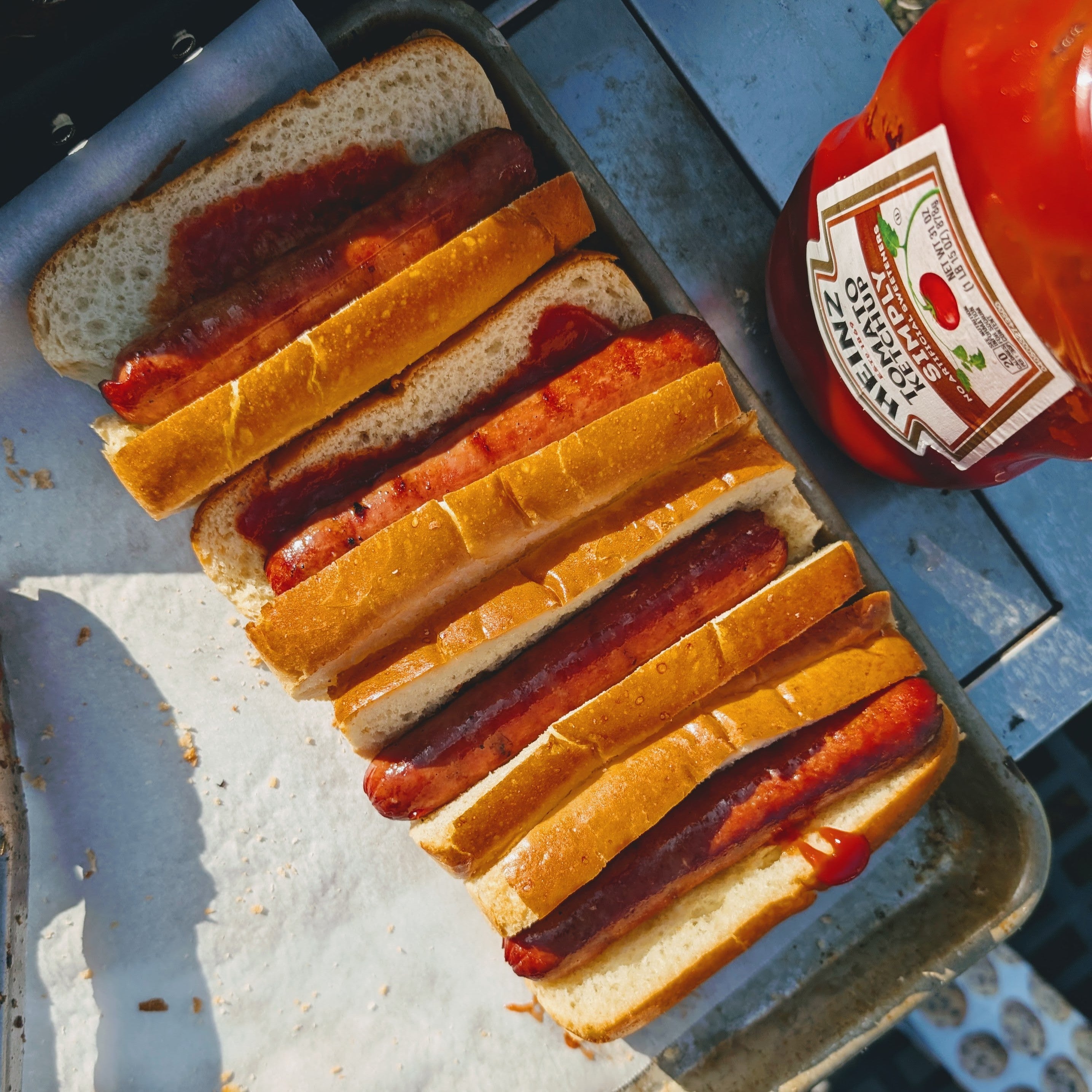 Four grilled hot dogs in buns on a tray, alongside a bottle labeled HEINZ TOMATO KETCHUP