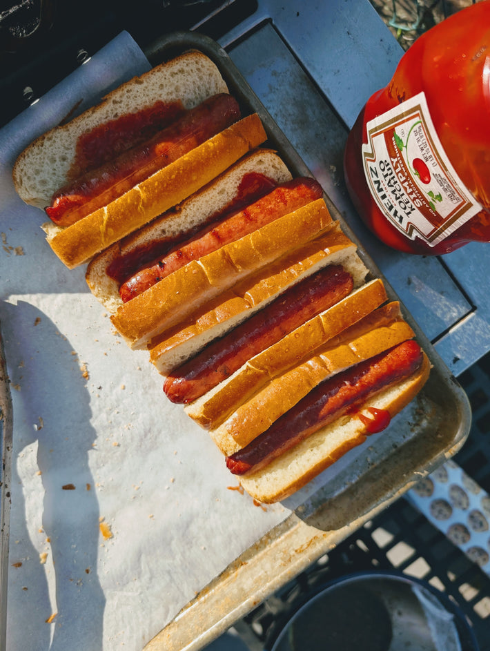 Grilled hot dogs in split buns on a tray, with a ketchup bottle showing label HEINZ TOMATO KETCHUP.